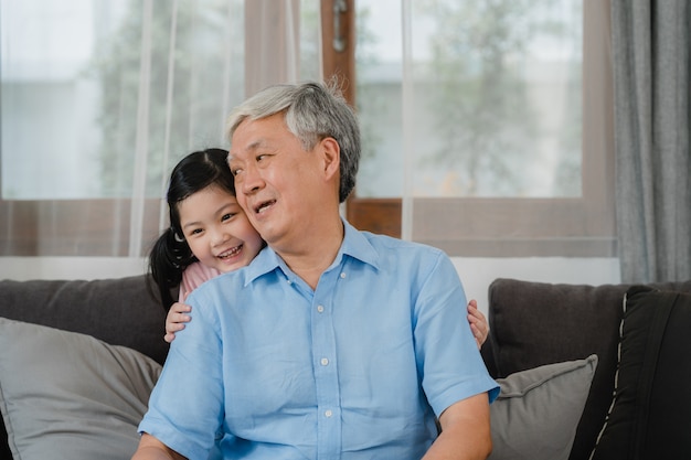 Free photo asian grandfather talking with granddaughter at home. senior chinese, grandpa happy relax with young granddaughter girl using family time relax with young girl kid lying on sofa in living room.