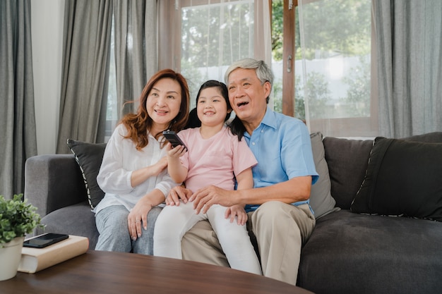 Free photo asian grandparents watch tv with granddaughter at home. senior chinese, grandfather and grandmother happy using family time relax with young girl kid lying on sofa in living room concept.