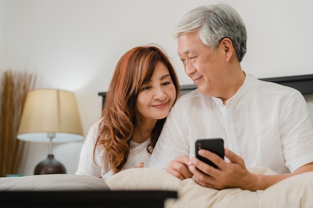 Free photo asian senior couple using mobile phone at home. asian senior chinese grandparents, husband and wife happy after wake up, watching movie lying on bed in bedroom at home in the morning concept.