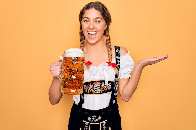 Beautiful blonde german woman with blue eyes wearing octoberfest dress drinking jar of beer very happy and excited winner expression celebrating victory screaming with big smile and raised hands