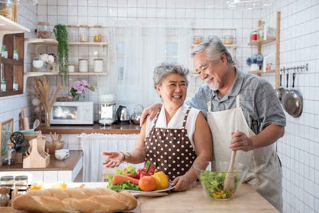 Photo senior couple having fun in kitchen with healthy food - retired people cooking meal at home with man and woman preparing lunch with bio vegetables - happy elderly concept with mature funny pensioner.