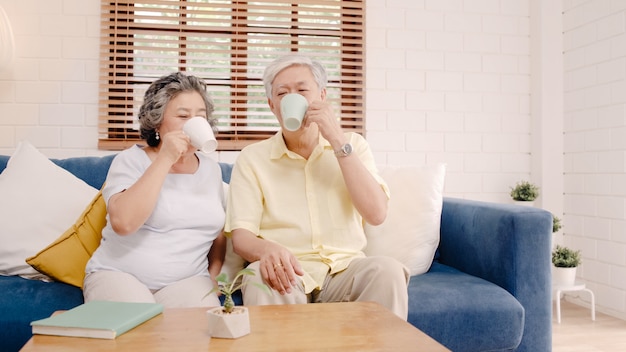 Free photo asian elderly couple drinking warm coffee and talking together in living room at home, couple enjoy love moment while lying on sofa when relaxed at home.