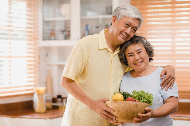 Free photo asian elderly couple feeling happy smiling and holding fruit and looking to camera while relax in kitchen at home.