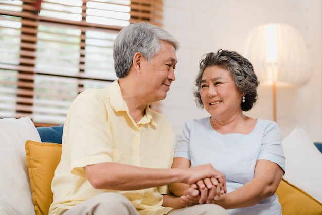 Free photo asian elderly couple holding their hands while taking together in living room, couple feeling happy share and support each other lying on sofa at home.