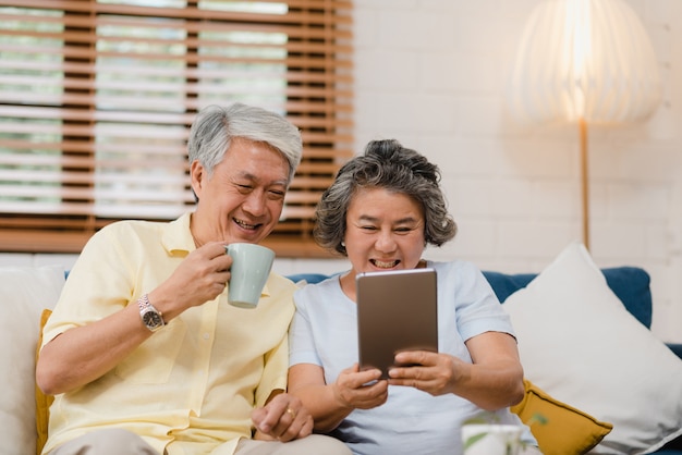 Free photo asian elderly couple using tablet and drinking coffee in living room at home, couple enjoy love moment while lying on sofa when relaxed at home.