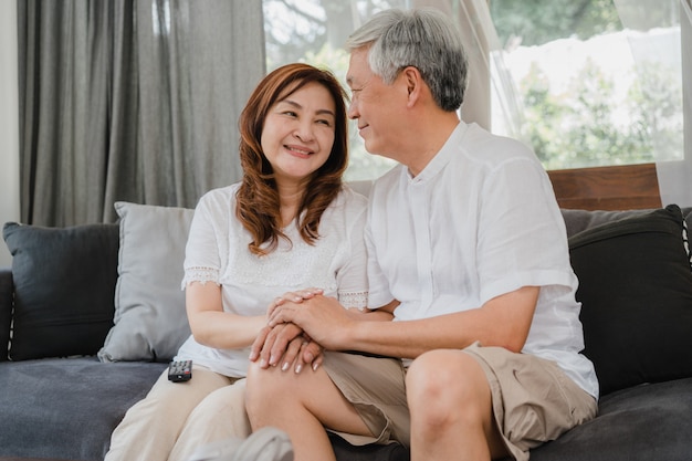 Free photo asian senior couple relax at home. asian senior chinese grandparents, husband and wife happy smile hug talking together while lying on sofa in living room at home concept.