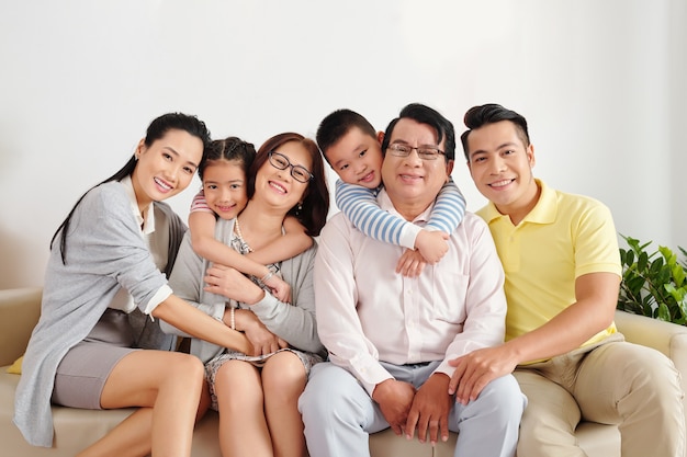 Photo cheerful vietnamese children, parents and grandparents sitting on sofa in living room, hugging and looking at camera