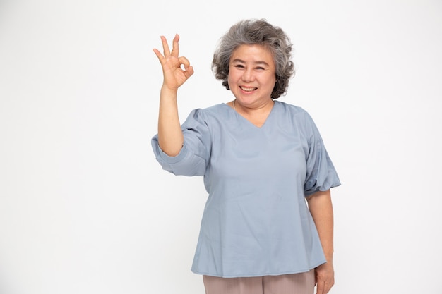 Photo portrait of happy senior asian woman gesture or showing hand ok and looking at camera isolated on white wall, older female feeling positive and enjoy concept